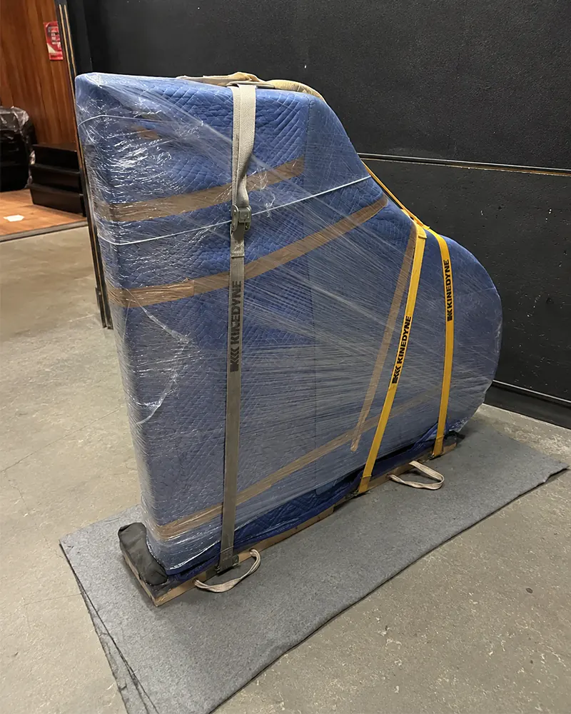 Upright piano wrapped in moving blankets, shrink wrap and straps on a piano board