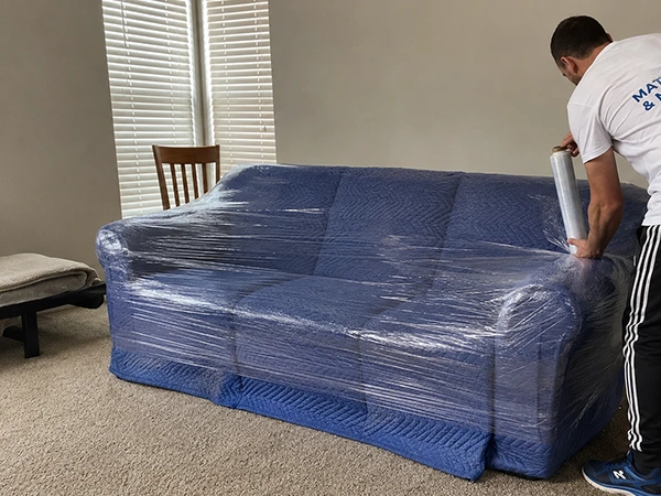 A mover shrink-wrapping a sofa that has already been padded with moving blankets