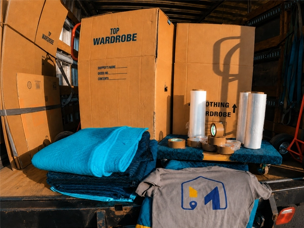 Moving blankets, rolls of stretch wrap, tape and a wardrobe carton loaded on the open tailgate of a moving truck
