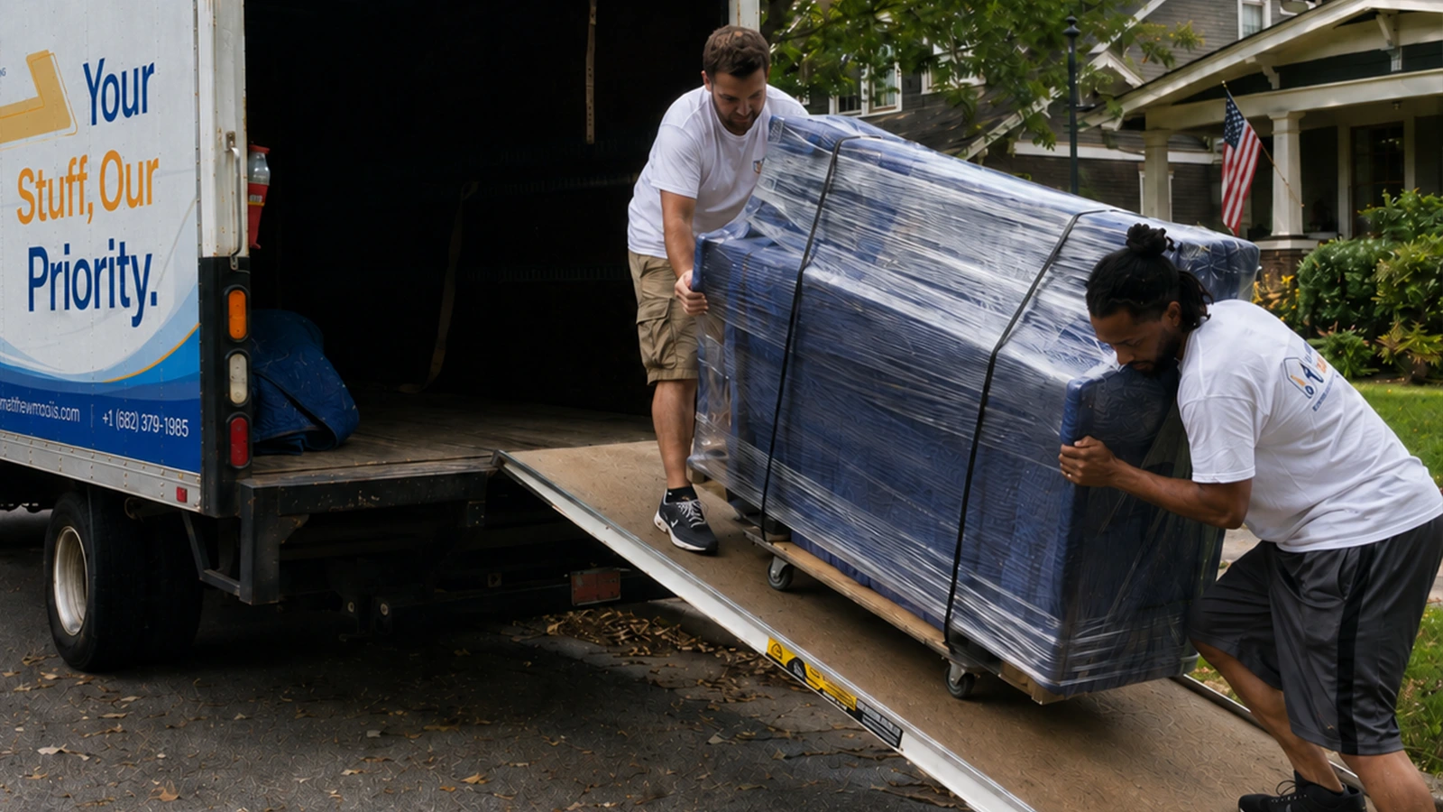 Two Matthew and Mois movers guiding a large shrink-wrapped piece up the ramp into a box truck on a residential street