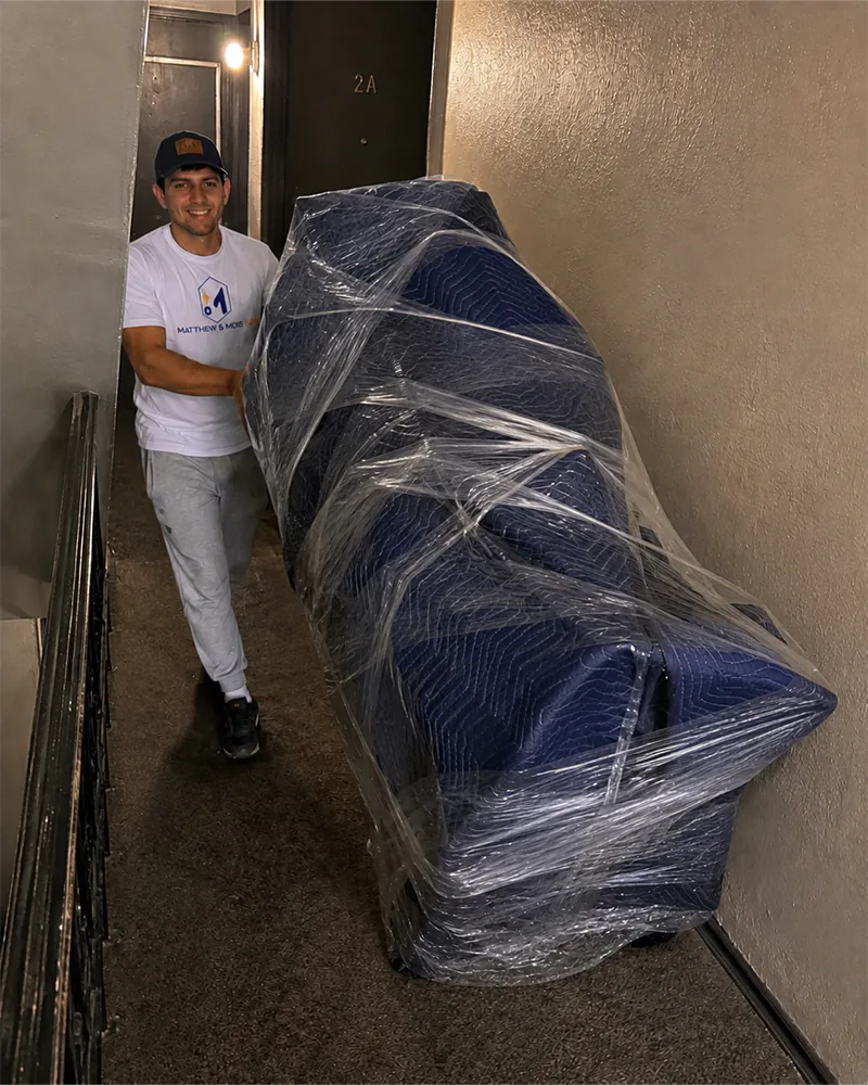 A Matthew and Mois mover easing a blanket-wrapped and shrink-wrapped sofa on a dolly through a narrow apartment corridor