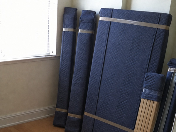 Bed rails, headboard panels and slats wrapped in moving blankets and taped, standing together against a bedroom wall